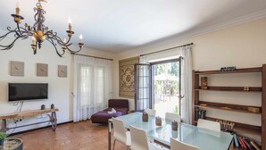 A dining area with a glass table, white chairs, a chandelier, a television, and a bookshelf with chess sets. - La Teja Holiday Home (Marchena)