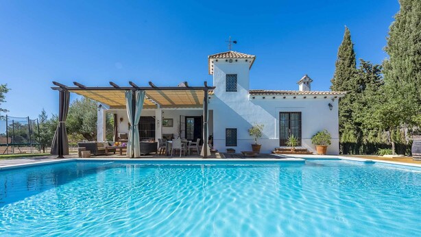 A poolside area with a pergola, outdoor seating, and a white building with a tiled roof. - La Teja Holiday Home (Marchena)