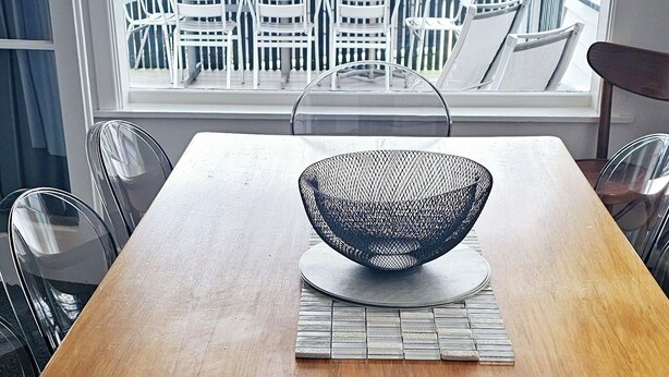 A dining area with a wooden table, white chairs, and a black mesh bowl centerpiece. A balcony with white chairs and a view of a body of water and distant hills is visible through the window.