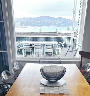 Een eetruimte met een houten tafel, witte stoelen en een zwarte net kuip als middelpunt. Door het raam is een balkon met witte stoelen en uitzicht op een waterpartij en heuvels zichtbaar.