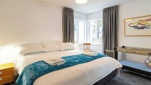 A hotel room with a bed, a nightstand, a chair, a window with curtains, and a painting on the wall.