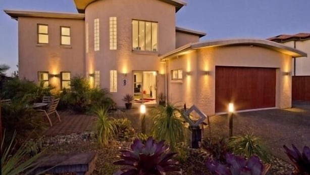 Une maison moderne à deux étages, dotée d’un toit aux lignes courbes, d’une allée couverte et d’un jardin aménagé avec diverses plantes et un espace de détente.