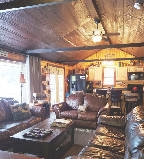 A cozy cabin interior with leather sofas, a wooden ceiling, and a kitchen area.