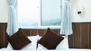 Ein Bett mit zwei braunen Kissen, einem weißen Kissen und Blick auf Berge durch ein Fenster mit Vorhängen.