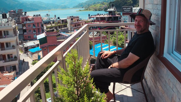 Ein Mann sitzt auf einem Balkon mit Blick auf einen See, im Hintergrund Gebäude und Berge.