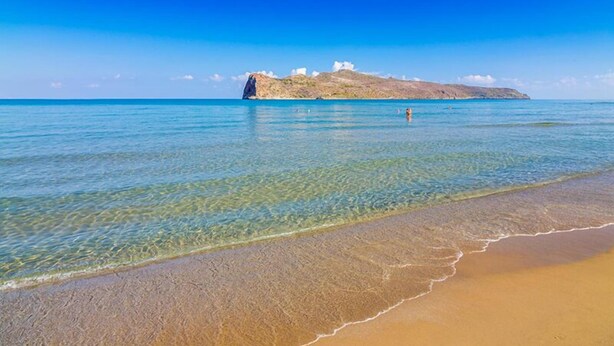 A beach with clear water, a sandy shore, and a small island with white buildings in the distance. - Rotunda Suites (Chania)