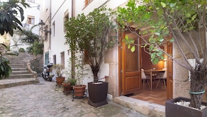 Un patio con suelo de piedra, plantas en macetas, una moto y una puerta de madera que da a una habitación con mesa y sillas.