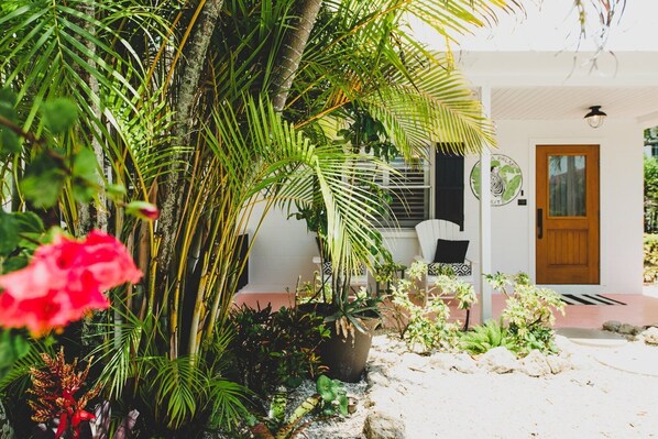 A tropical garden leading to a white building with a wooden door and a white chair outside.