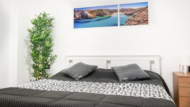 Ett sovrum med en säng, en växt placerad på sängbordet, ett sängbord och en tavla med ett kustlandskap som hänger på väggen.