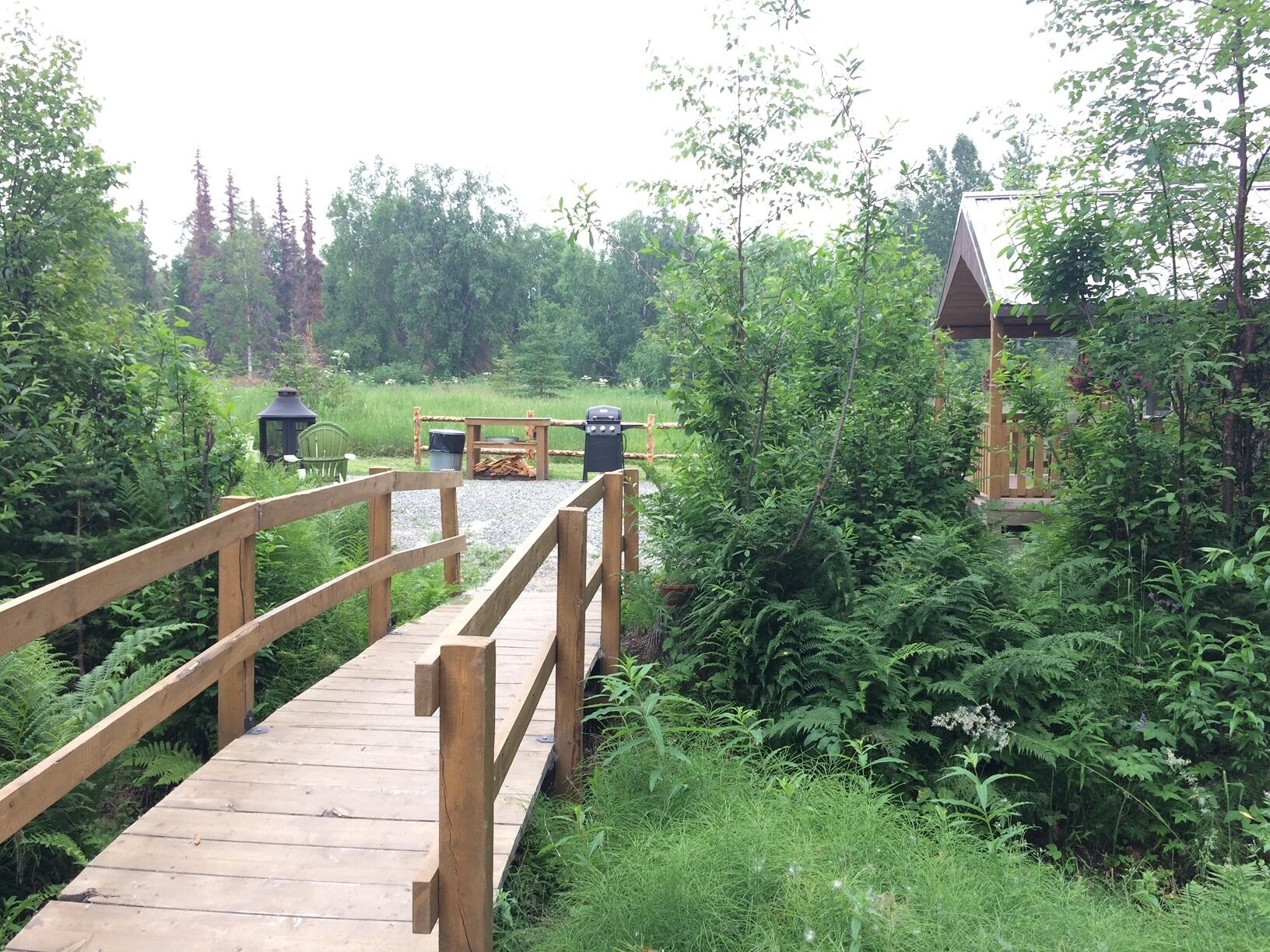Eine Holzbrücke zu einem rustikalen Außenbereich mit Grill und Laterne.