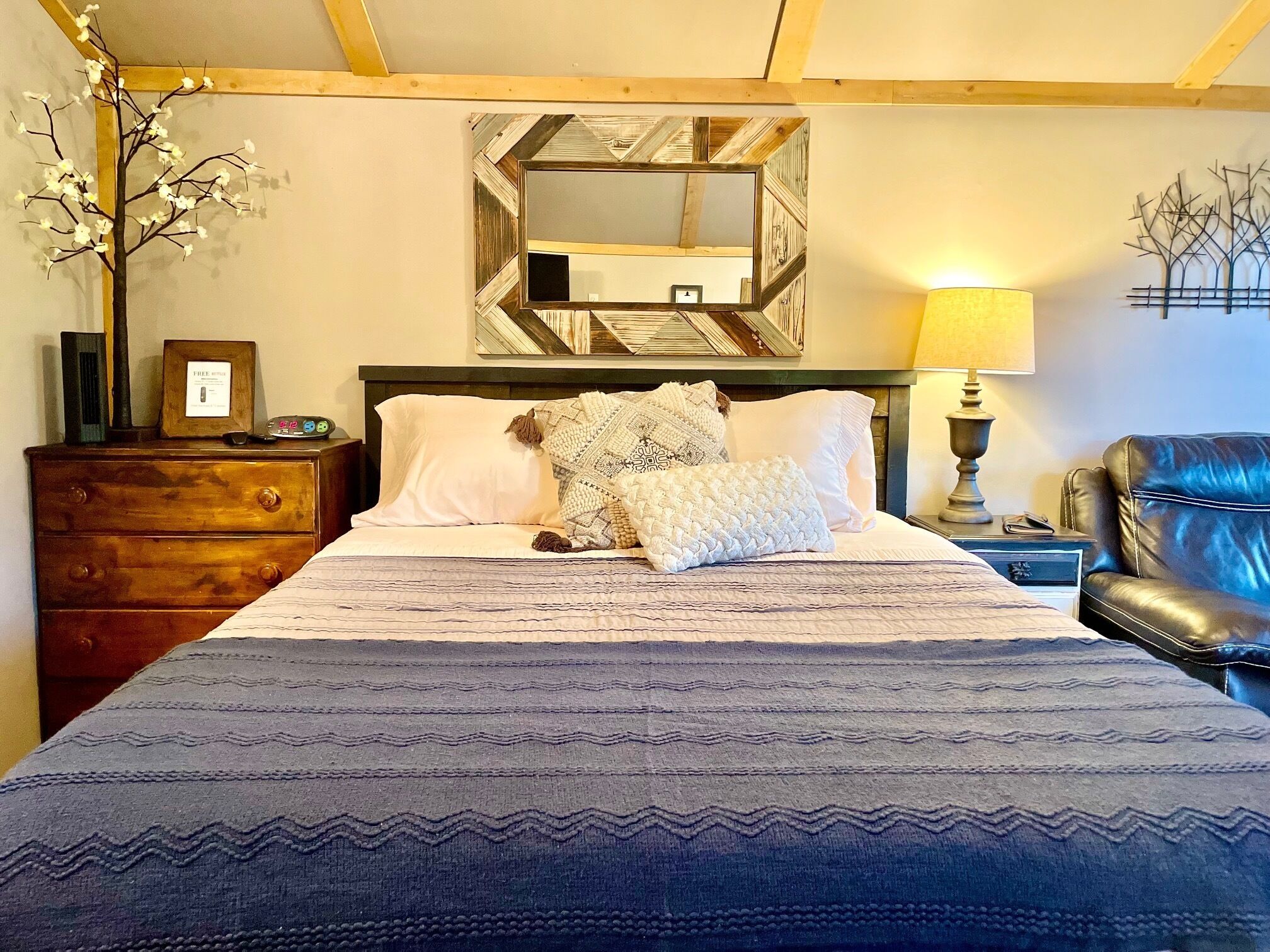 Een opgemaakt bed met een geborduurd beddekleed, een houten dressoir met spiegel, een lamp en een stoel.
