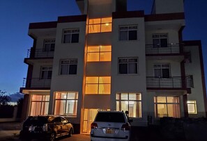 A multi-story residential building with a central entrance, two cars parked in front, and multiple windows with warm lighting. - Ambiere House (Mtwara)