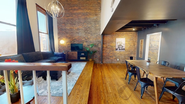 Una sala de estar moderna con pared de ladrillo, un sofá, una mesa de comedor de madera con sillas y un televisor sobre un mueble.