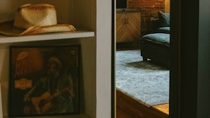 Una habitación con pared de ladrillo, suelo de madera, cama, estante con sombreros y un cuadro enmarcado.