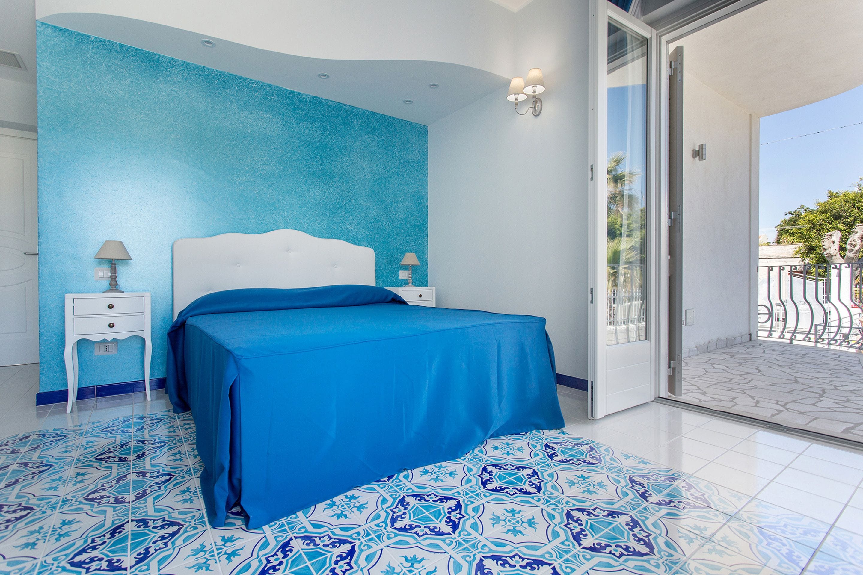 A bedroom with a blue bedspread, a white bedside table with a lamp, and a patterned floor.