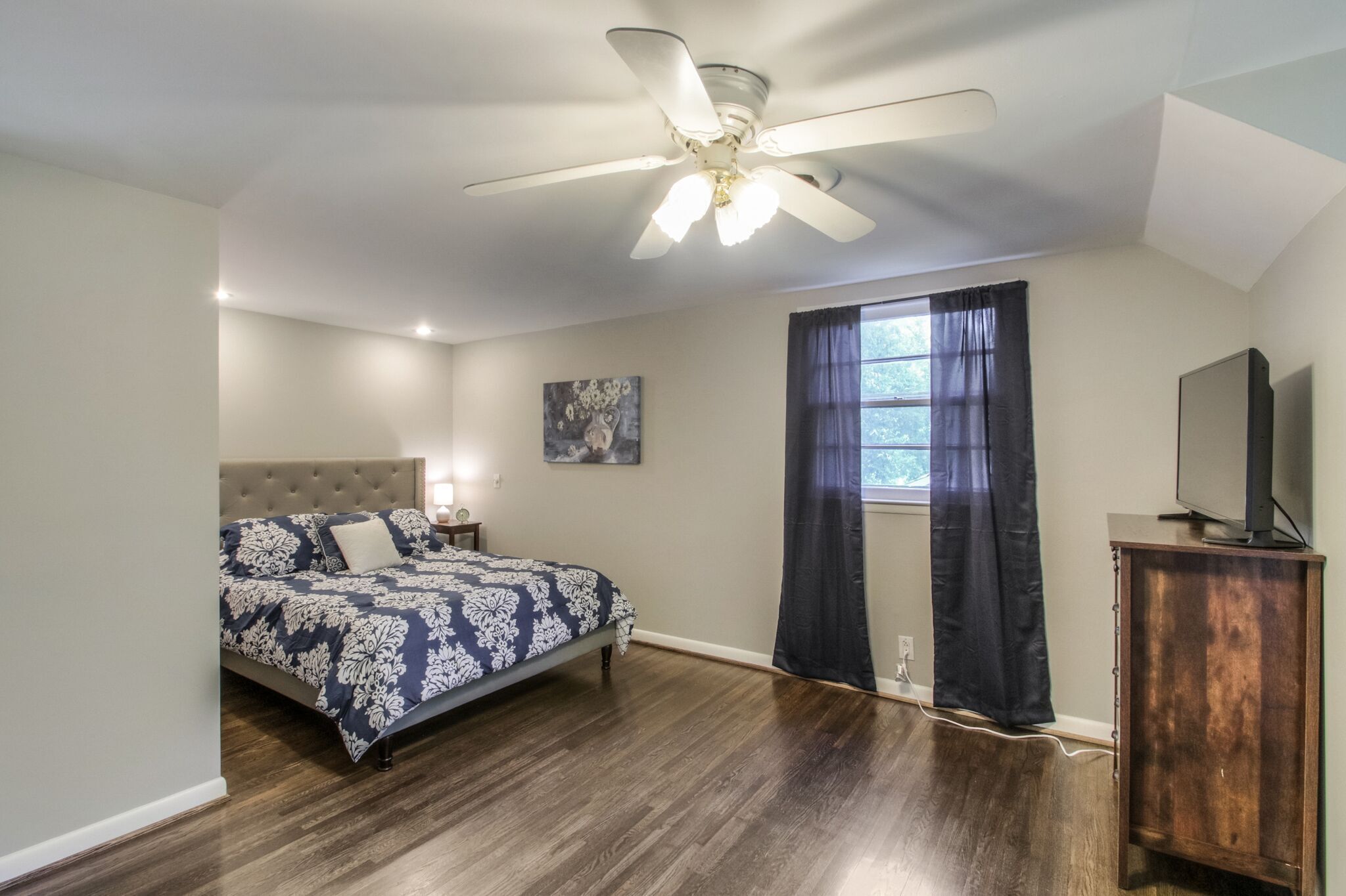 Une chambre à coucher avec un lit, un ventilateur de plafond, un téléviseur posé sur un meuble en bois et une fenêtre avec des rideaux.