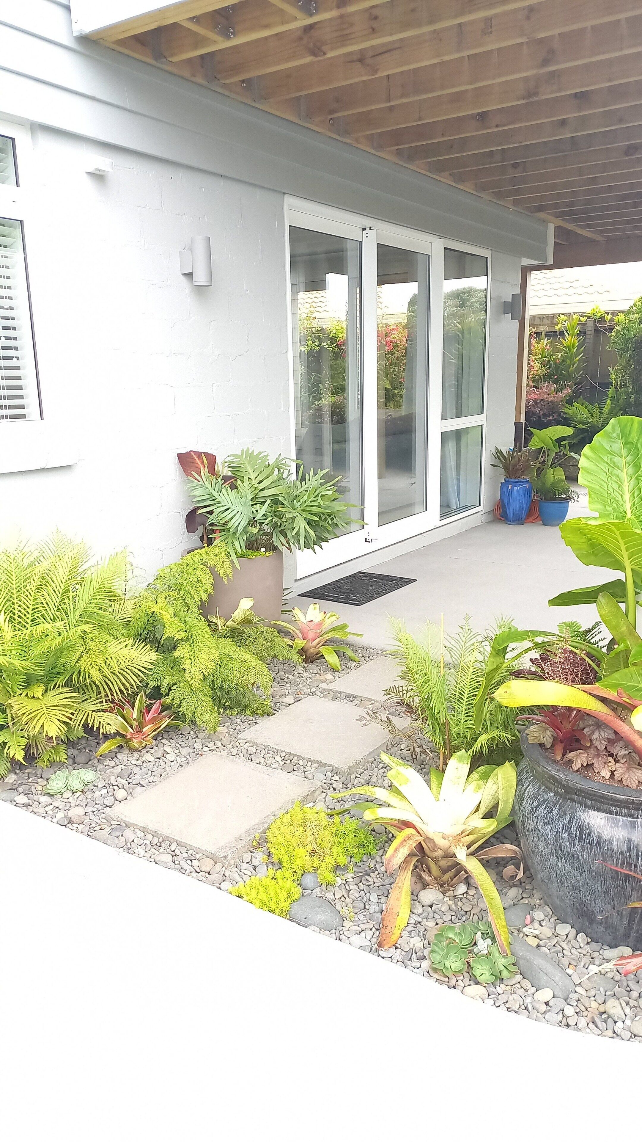 Un patio couvert avec un chemin en béton, des plantes en pot et une porte-fenêtre coulissante donnant sur un jardin.