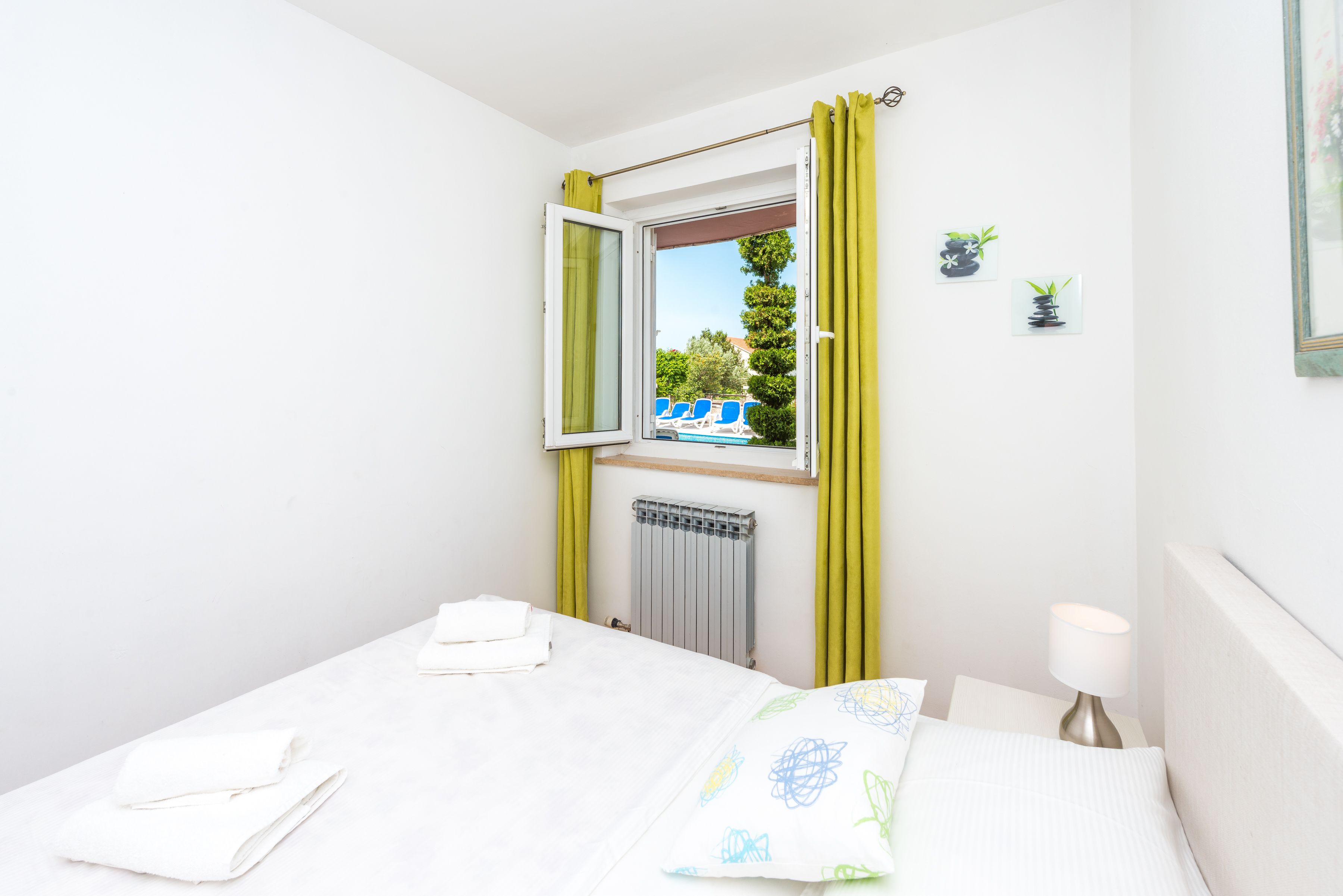 A bedroom with a bed, a radiator, a window with curtains, a bedside table with a lamp, and a view of a pool and chairs outside.