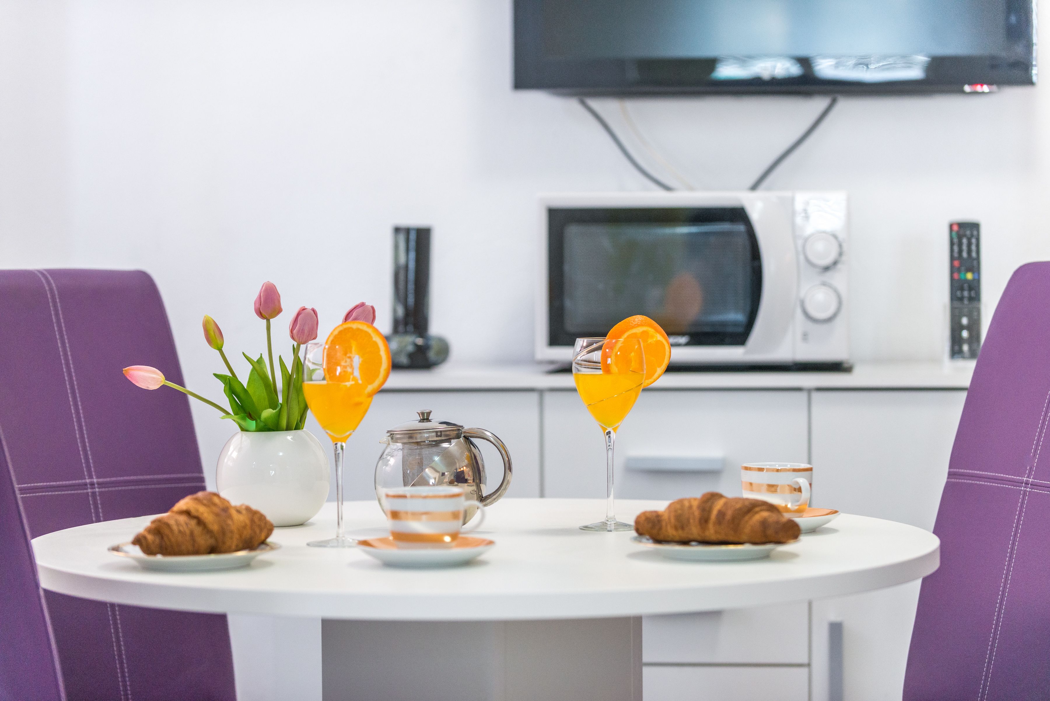 Una mesa de desayuno con cruasanes, un jarrón con tulipanes, una tetera y dos vasos de jugo de naranja.