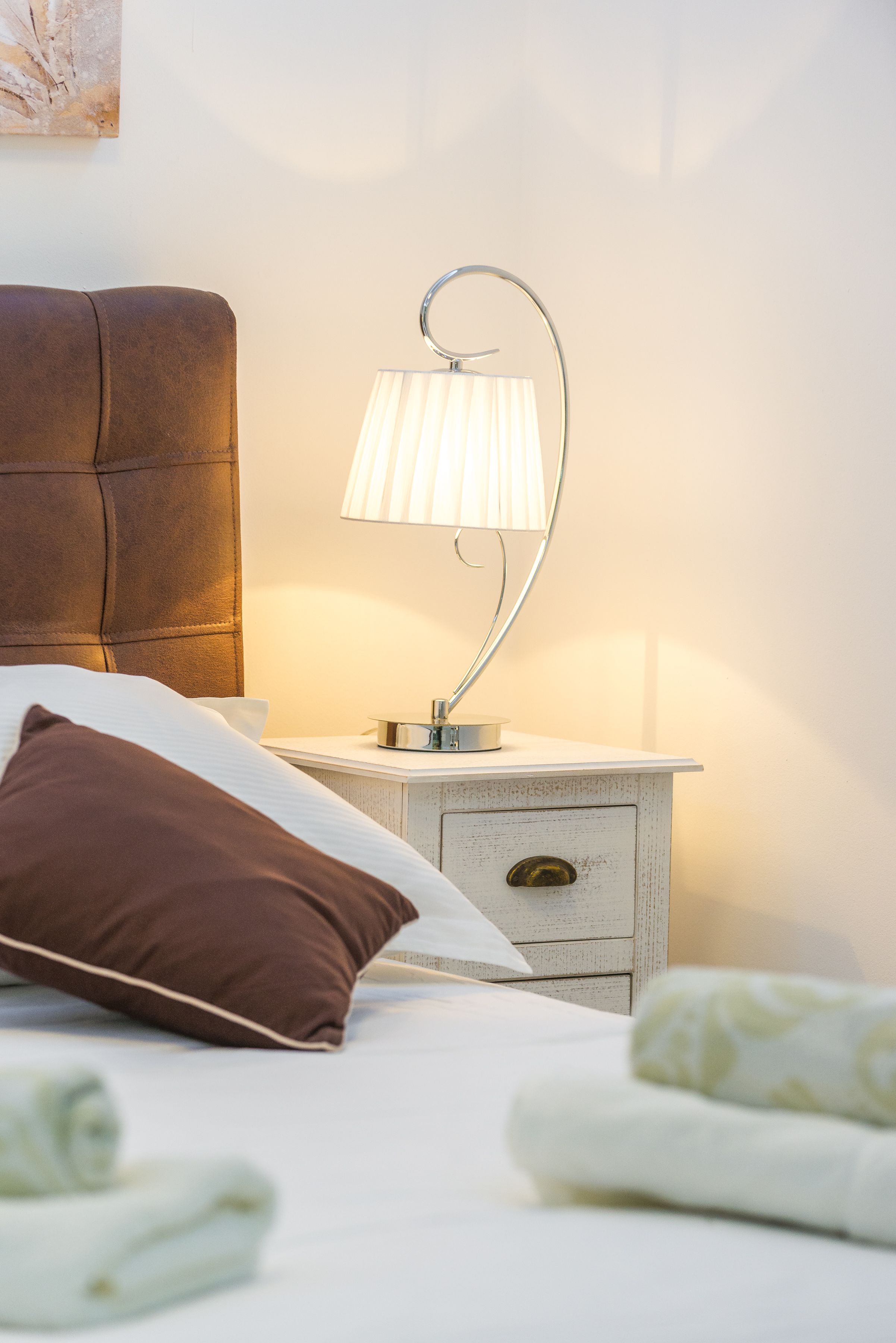 A neatly made bed with a brown pillow, a white pillow, and a white blanket. There is a bedside table with a lamp and a small drawer.