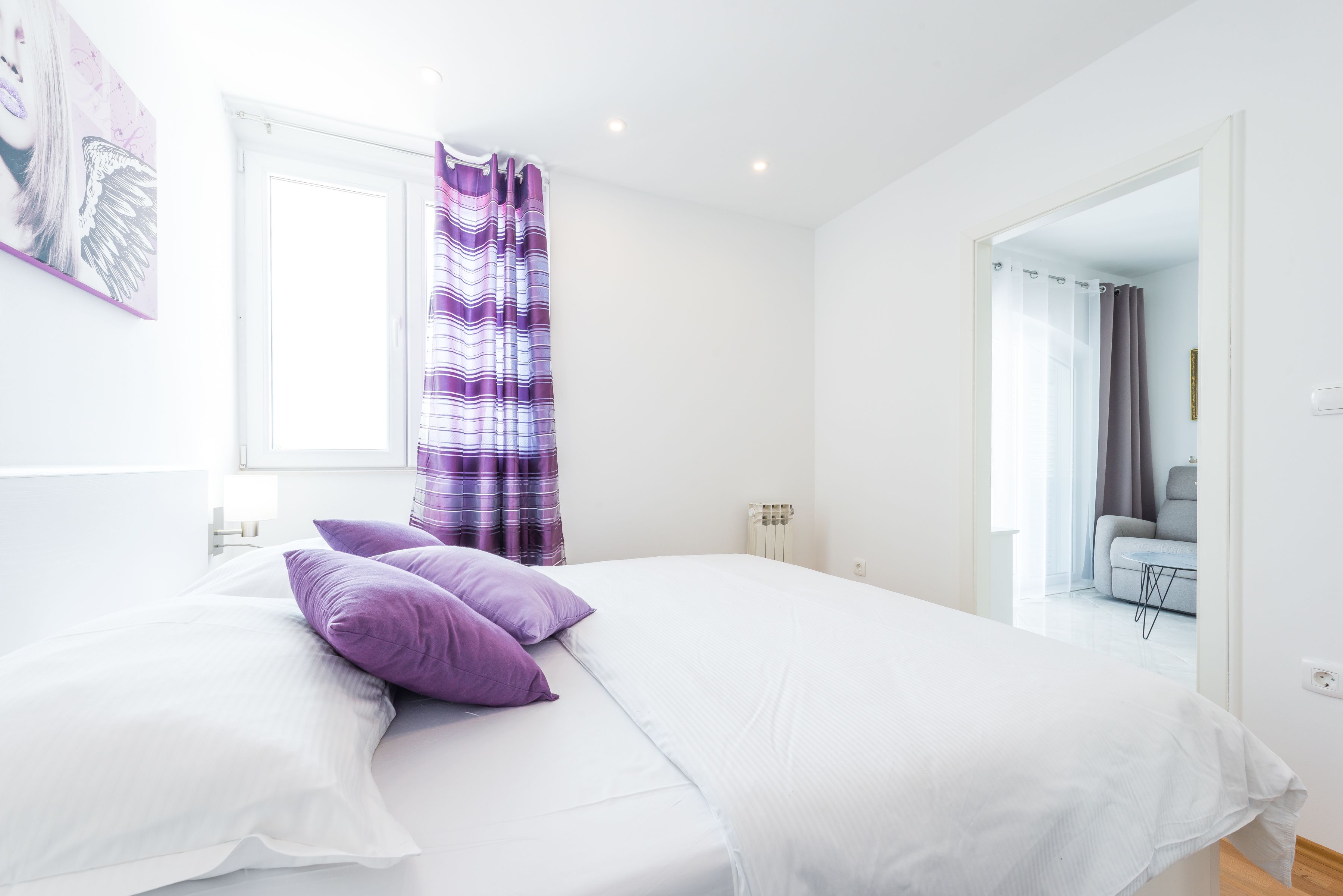 Un dormitorio con cama, almohadas moradas, una ventana con cortinas y vistas a una sala de estar.