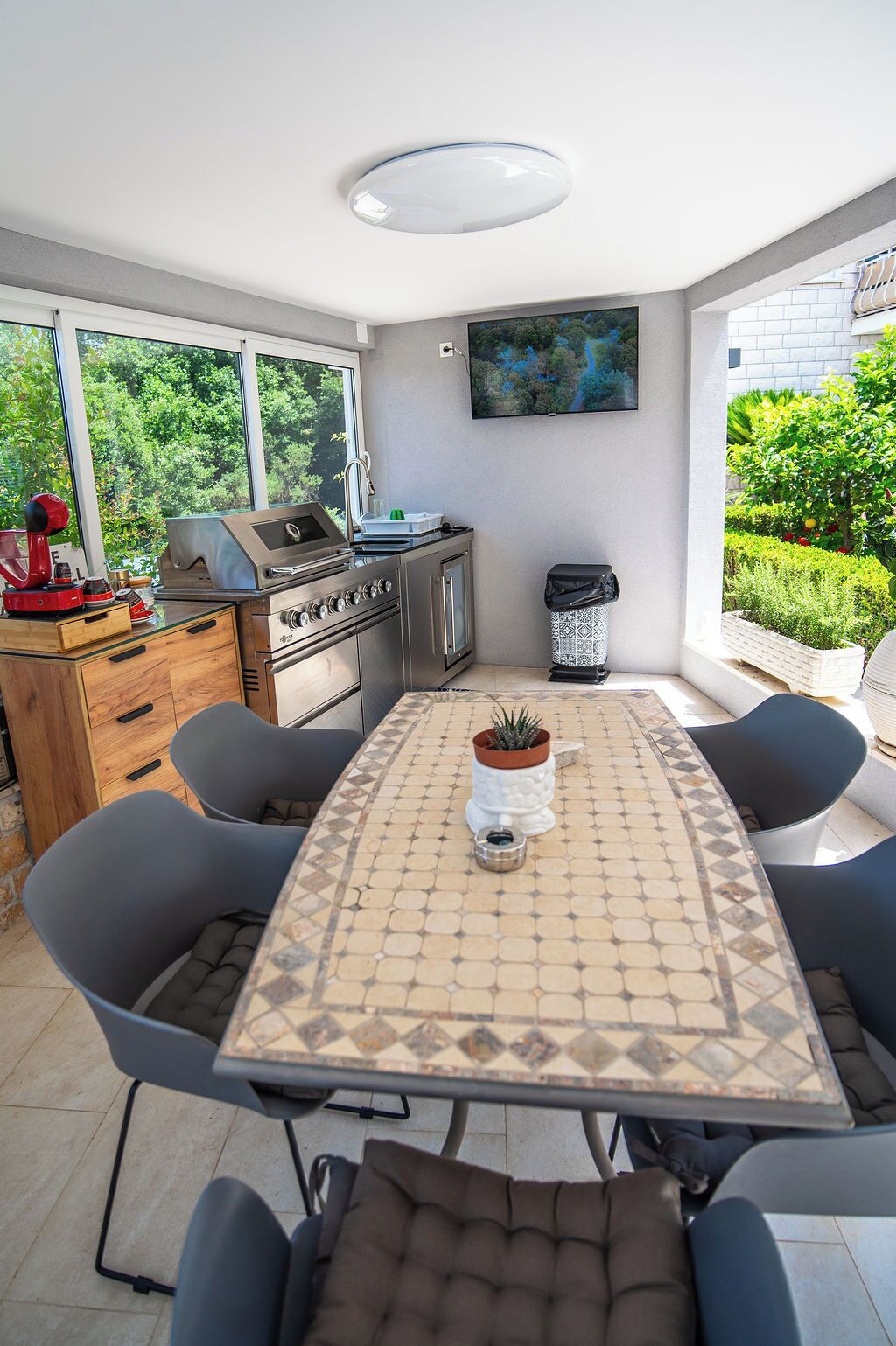 A modern outdoor kitchen with a grill, dining table, and chairs, surrounded by large windows and a garden view.