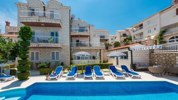 A pool area with blue lounge chairs and umbrellas, surrounded by multi-story buildings with balconies and a stone wall.