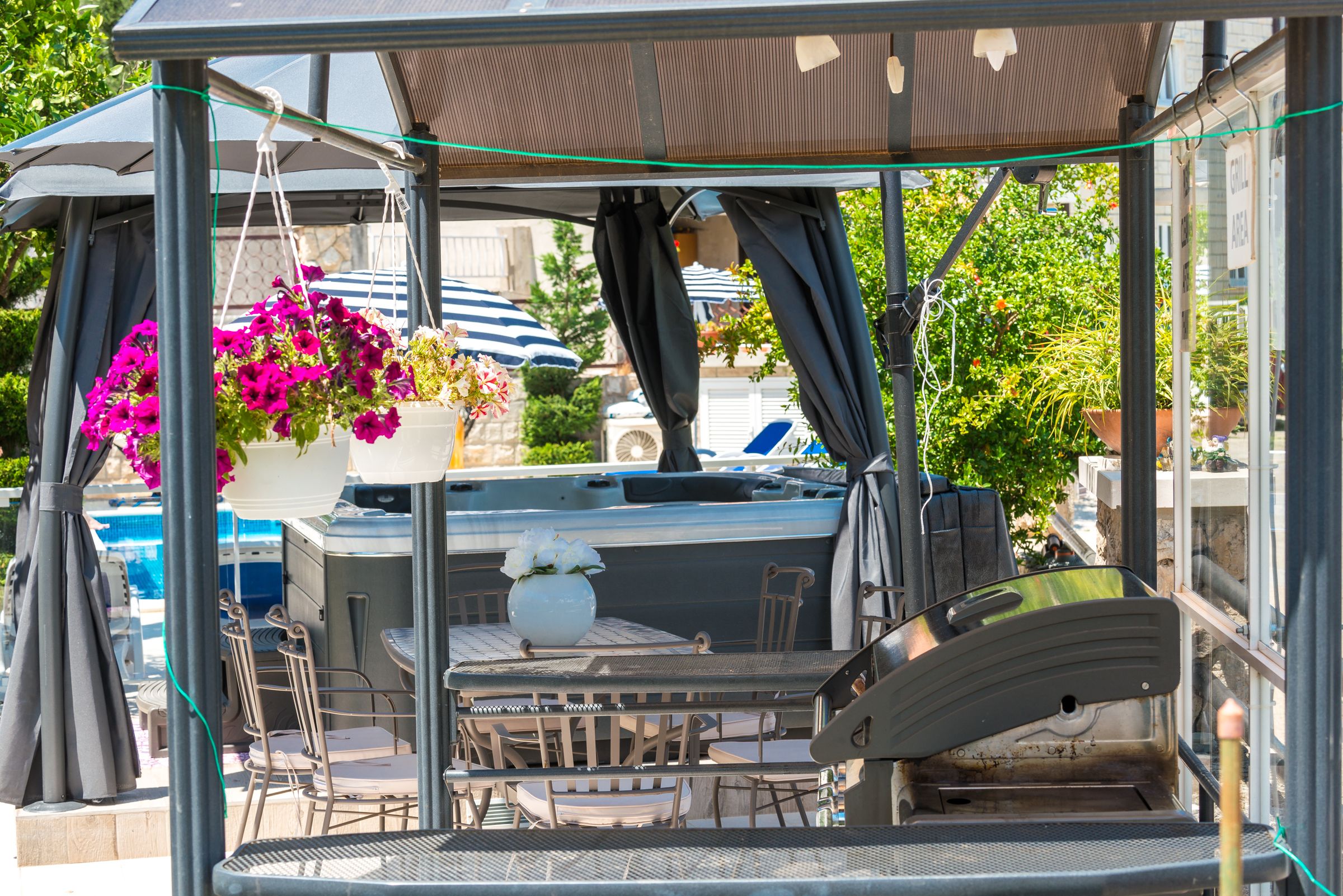 A covered outdoor seating area with a table and chairs, a grill, and hanging flowers.