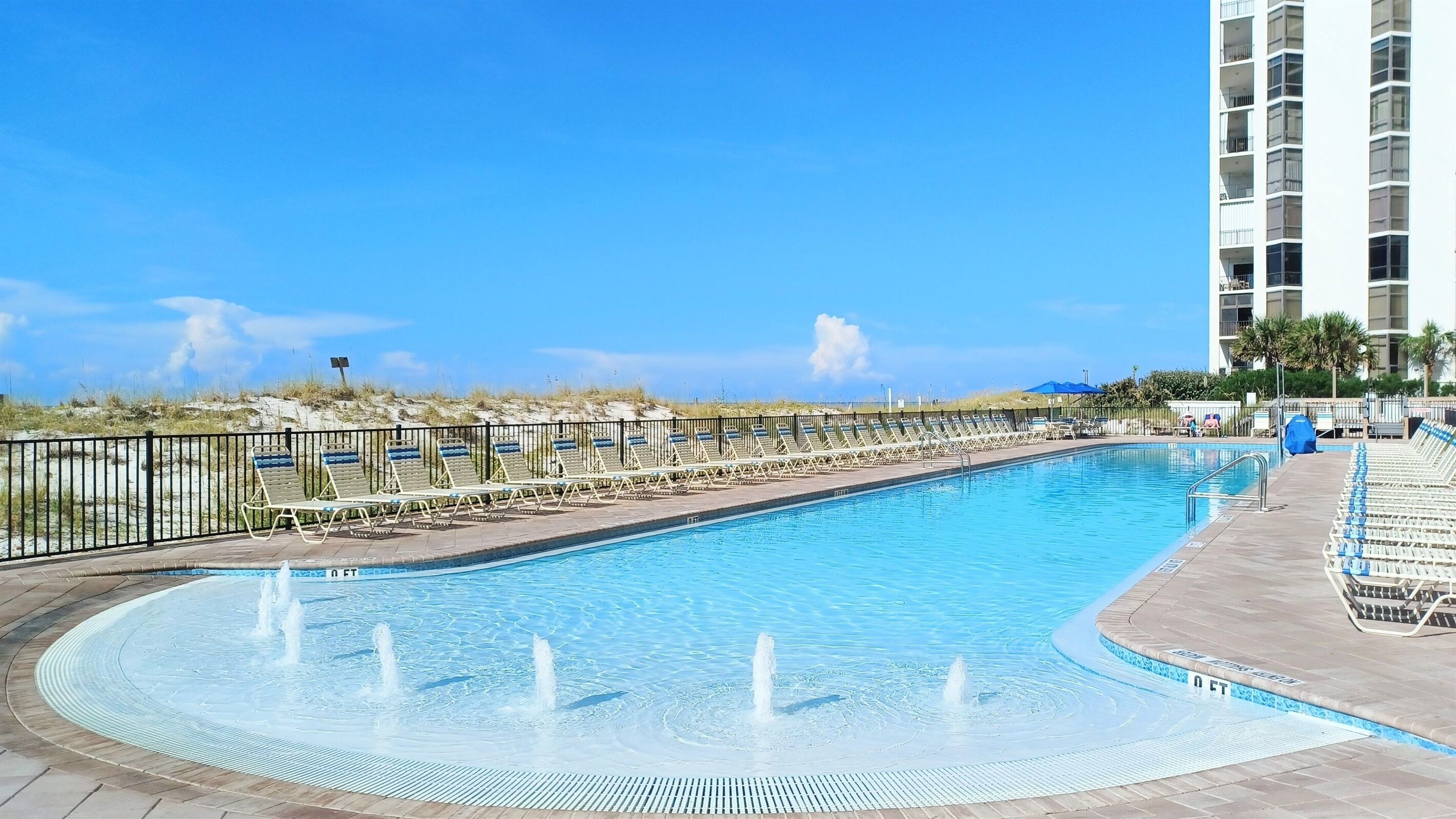 A swimming pool with a fountain feature, surrounded by lounge chairs and a high-rise building in the background.