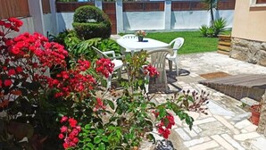 A patio with a white table and chairs, surrounded by red flowers and greenery. - Jardim da Maresia was renovated for your convenience and unforgettable holidays. (São João das Lampas)