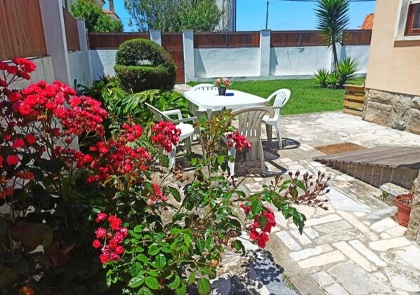 Un patio con una mesa y sillas blancas, rodeado de flores rojas y follaje.
