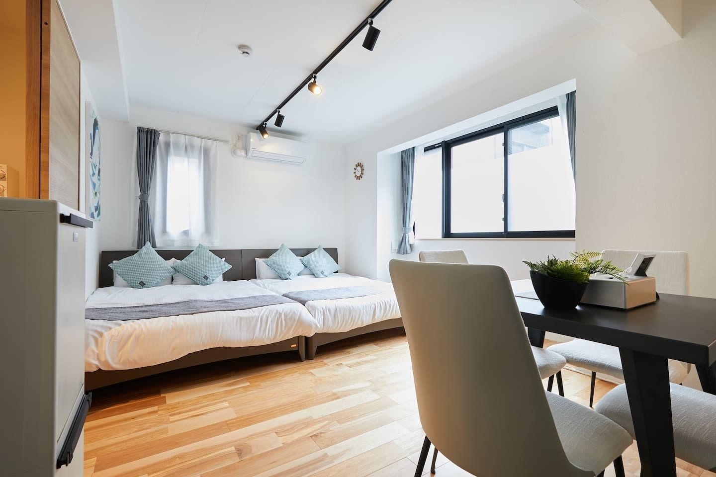 A modern bedroom with a bed, a desk, and a chair. There is a window providing natural light and a clock on the wall.