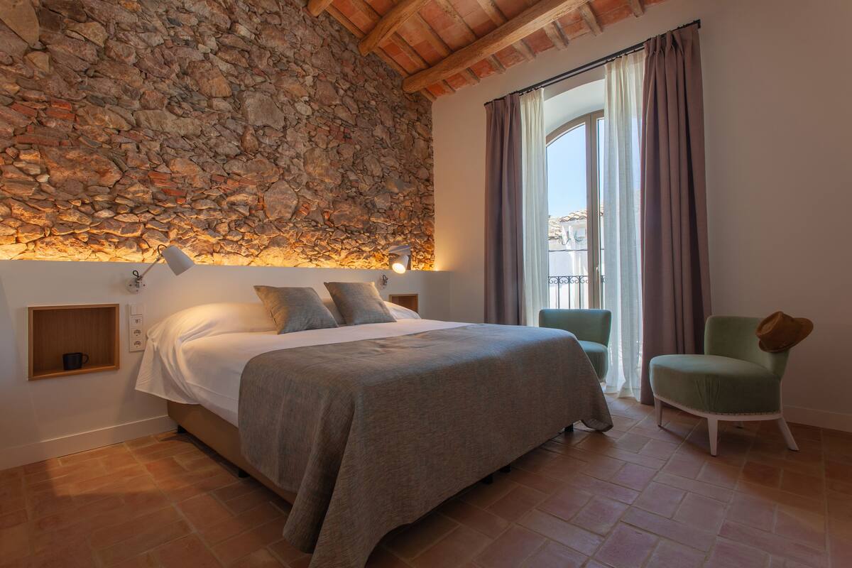 A bedroom with a stone wall, a bed with pillows, a chair, and a window with curtains.