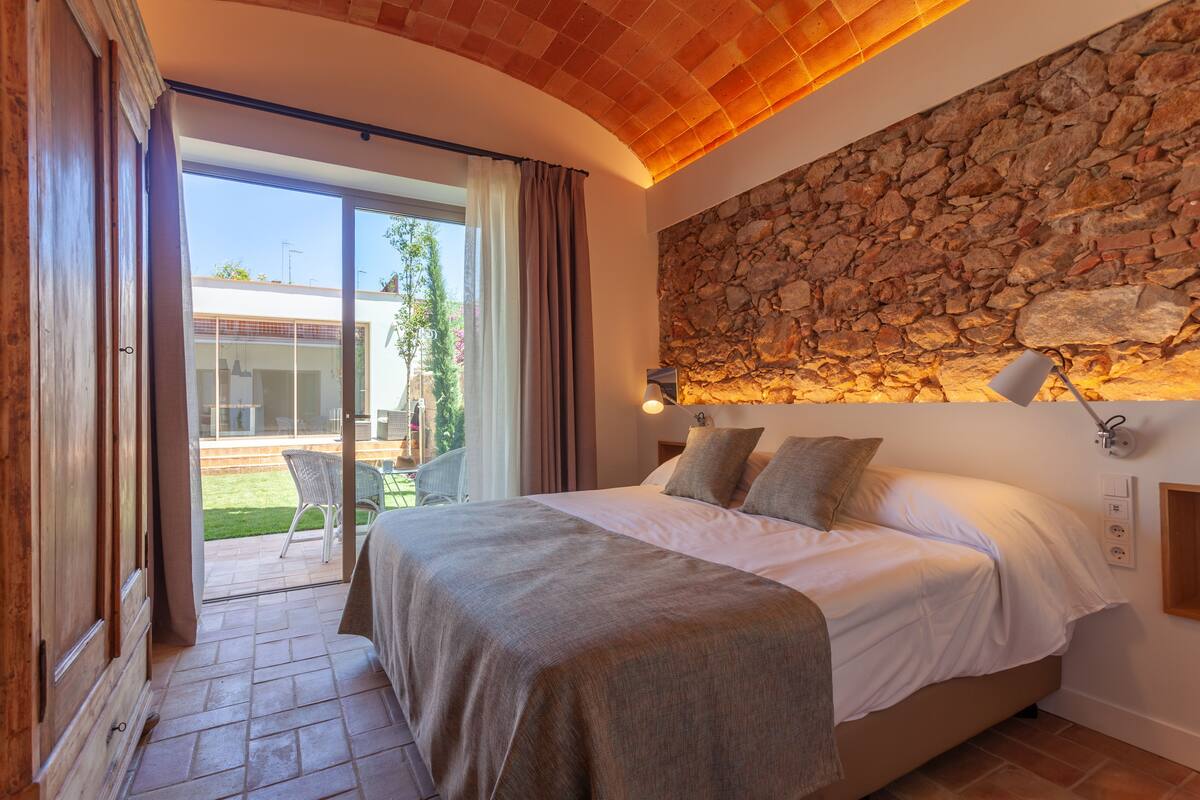 A bedroom with a stone wall, a bed with pillows, a nightstand, and a lamp.