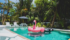 Une piscine agrémentée d’un flotteur en forme de flamant rose, entourée de palmiers et de chaises longues blanches.