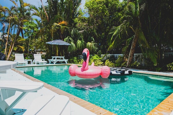 Une piscine agrémentée d’un flotteur en forme de flamant rose, entourée de palmiers et de chaises longues blanches.