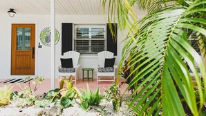 Un bâtiment blanc avec une porte en bois, deux chaises blanches avec des coussins noirs, un petit tabouret vert et une fenêtre avec des stores.