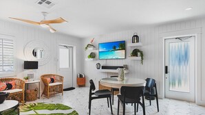 Un salon moderne avec une table de salle à manger ronde, des chaises noires, des canapés en osier, un ventilateur de plafond, un téléviseur à écran plat et une grande fenêtre donnant sur la plage.