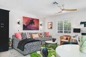 Une pièce avec un canapé à motifs zébrés, une table ronde, un ventilateur de plafond, une fenêtre avec des volets et un tableau encadré accroché au mur.