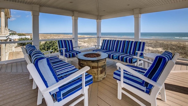 A covered outdoor seating area with white chairs and blue-striped cushions, a stone fire pit, and a view of the beach and ocean.