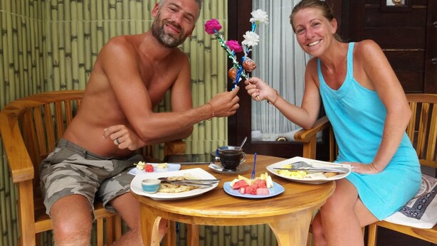 Två personer sitter vid ett träbord med tallrikar fyllda med mat, en kopp och en vas med blommor.