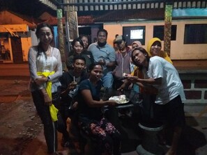 A group of people gathered around a table with a cake, in an outdoor setting with a building in the background. - Citiz's House (Penida Island)