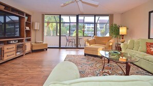 A spacious living room with a large window, a ceiling fan, and a green couch. - Spacious Condo in a Great Location! Private Beach, 2 Pools, 2 Jaccuzi, Fishing Deck & More! (Siesta Key)