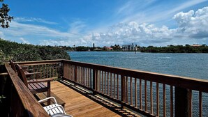 A wooden deck with a railing overlooking a body of water, with buildings and greenery in the background. - Spacious Condo in a Great Location! Private Beach, 2 Pools, 2 Jaccuzi, Fishing Deck & More! (Siesta Key)