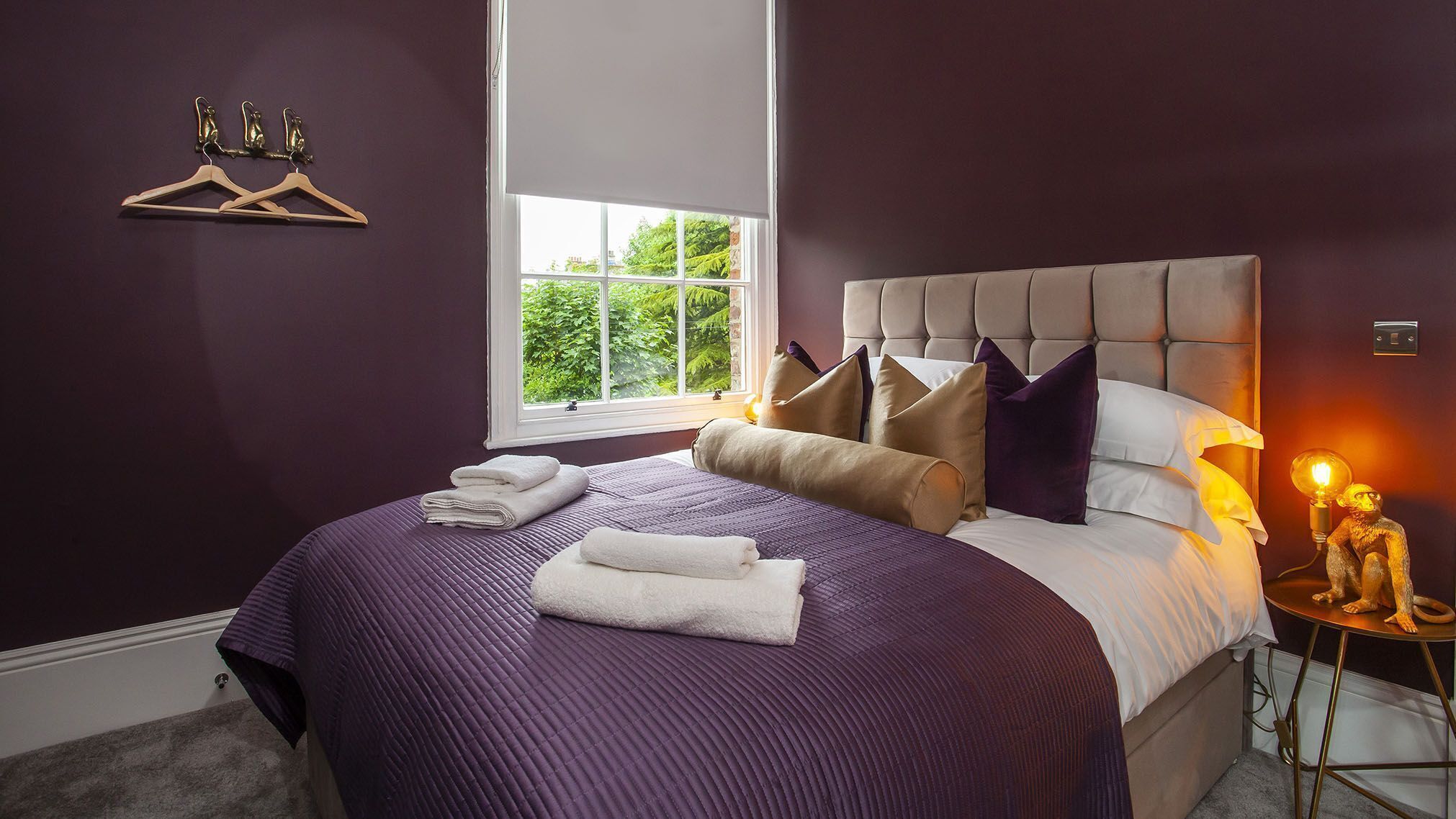 Une chambre avec un grand lit, une fenêtre donnant sur un paysage verdoyant, une table de chevet avec une lampe et un porte-manteau fixé au mur.
