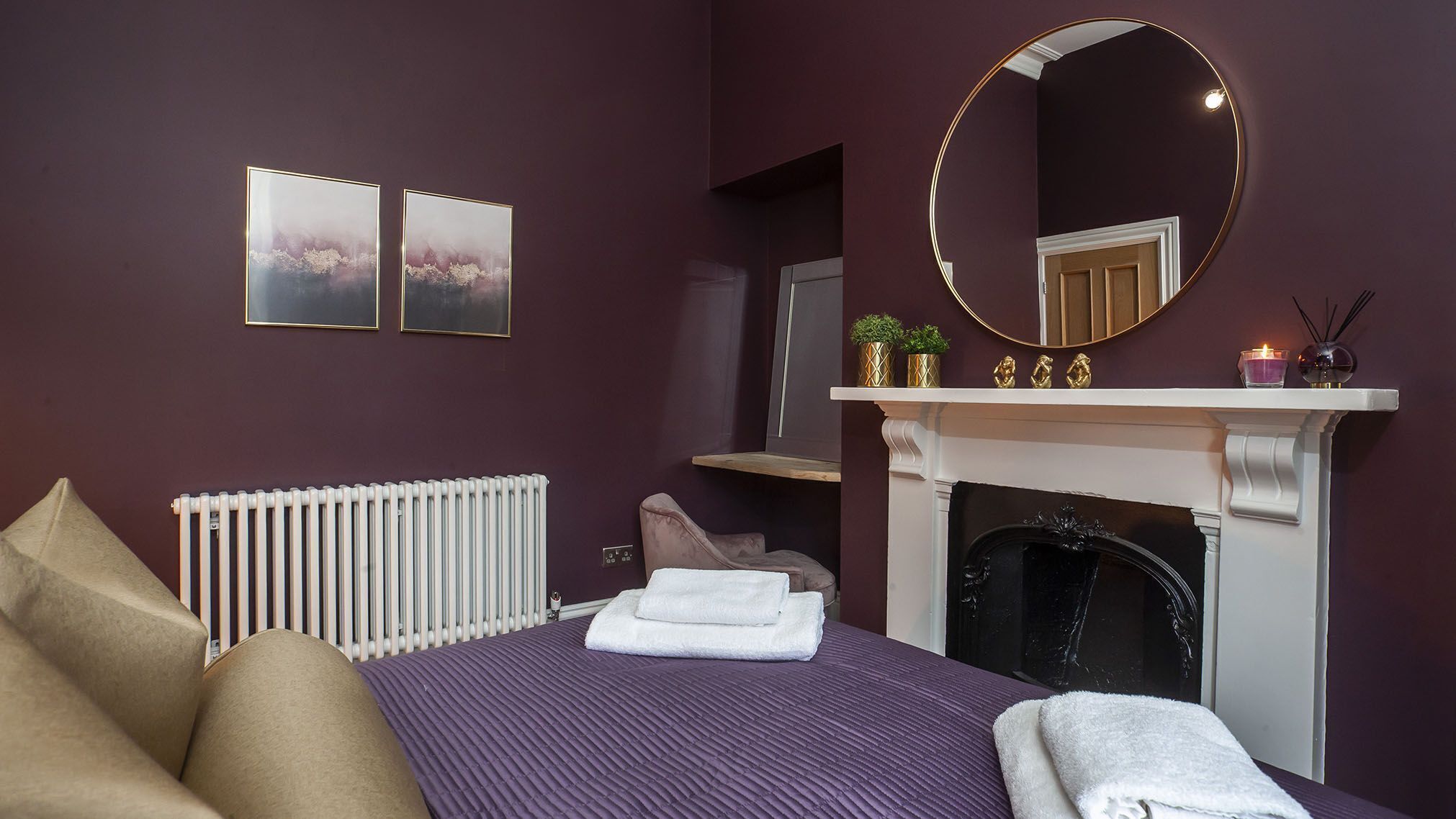 A bedroom with a purple bedspread, a white radiator, a fireplace, and two framed pictures on the wall.