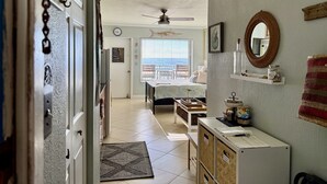 A hallway leading to a bedroom with a bed, a desk, and a chair. There is a ceiling fan, a window with blinds, and a door with a sign.