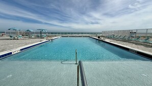 A rectangular pool with clear blue water, surrounded by lounge chairs and umbrellas, with a view of the ocean in the background.