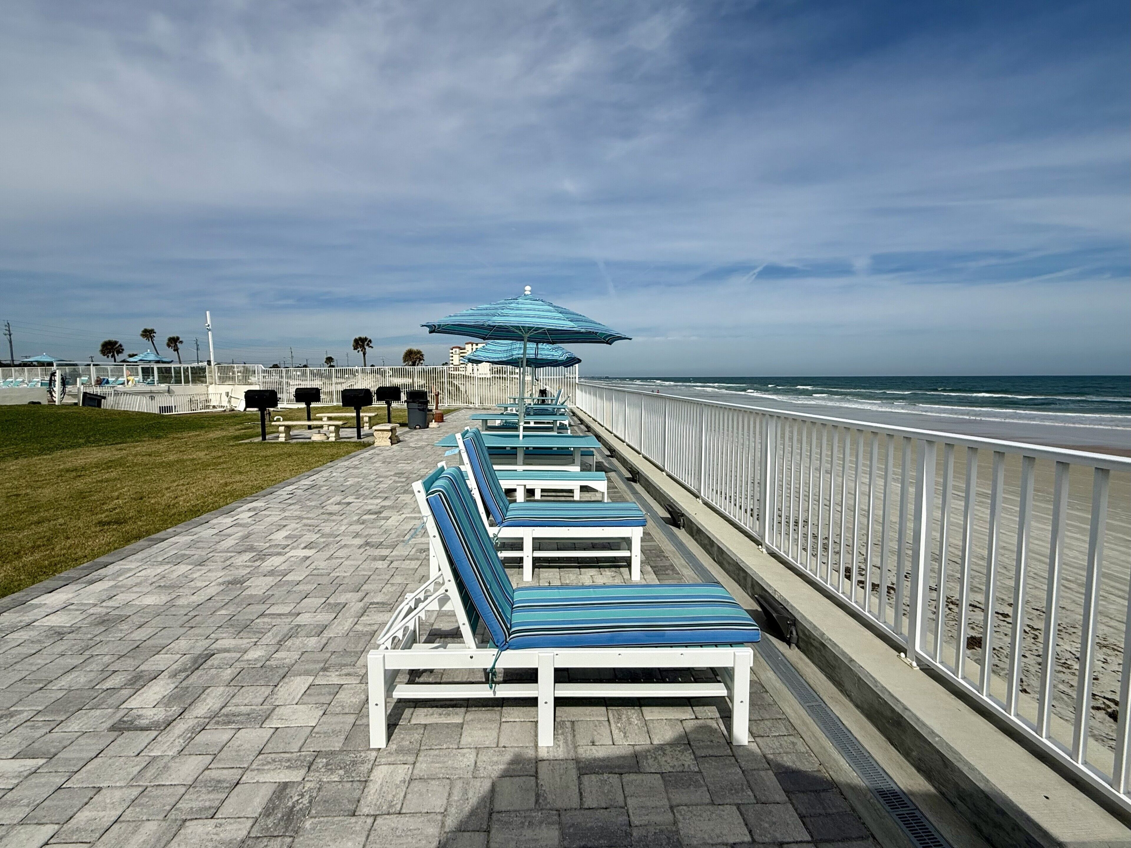 A beachside walkway with lounge chairs and umbrellas, a white fence, and a view of the ocean.