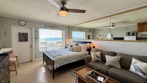 A bedroom with a bed, a bedside table, a dresser, a window with blinds, a ceiling fan, and a view of the ocean.